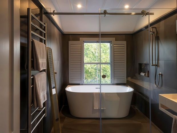 A refined contemporary bathroom with a freestanding white bathtub positioned beneath a timber-framed window with louvered shutters. A frameless glass shower enclosure occupies the right side, while a chrome heated towel rail with folded towels is visible on the left. Dark wall and floor finishes, recessed lighting, and streamlined chrome fixtures create a warm, moody atmosphere.
