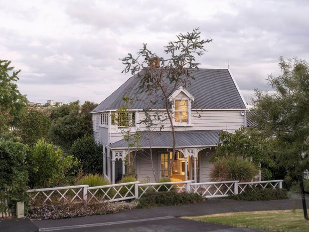 Exterior view of a two-storey heritage-style timber house with a steep corrugated metal gable roof, pale weatherboard cladding, decorative verandah detailing, and a white criss-cross front fence. The home is surrounded by established garden planting on a suburban street.