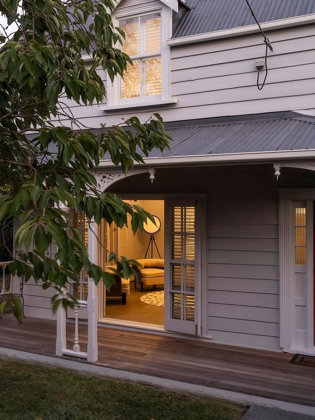 Exterior view of a refined timber-clad, two-storey house at dusk. The facade features horizontal weatherboards, a dark pitched metal roof, white-trimmed windows and doors, a covered veranda with decorative posts and corbels, and a timber deck. Warm interior lighting reveals a living room through open glazed doors, while leafy vegetation partially frames the house.