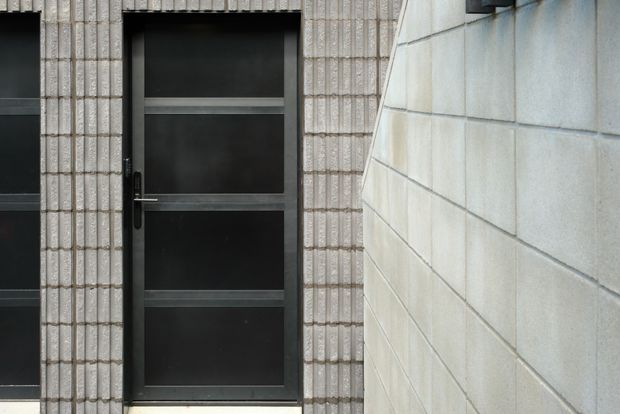 Close-up exterior architectural view of a dark metal-framed entry door with four opaque dark panels, set within a vertically textured light brick or tile facade. A pale smooth block wall forms the right side of a narrow passage, creating a restrained, contemporary composition with strong horizontal and vertical lines.
