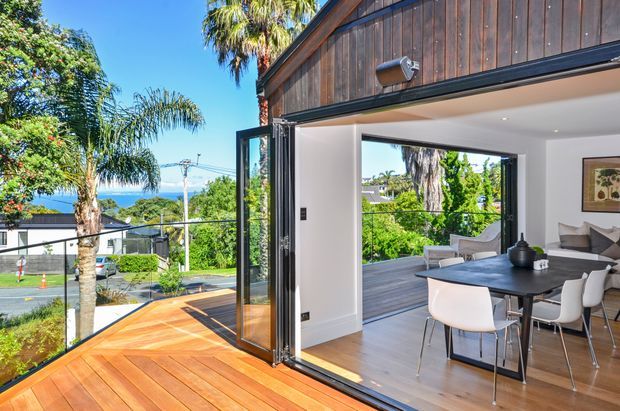 Bright contemporary open-plan dining and living area opening onto a timber deck through fully glazed folding doors. The room features white walls, timber flooring, a dark dining table with white chairs, and a warm timber-clad upper exterior facade. Palm trees, greenery, and a distant coastal or urban outlook are visible outside.