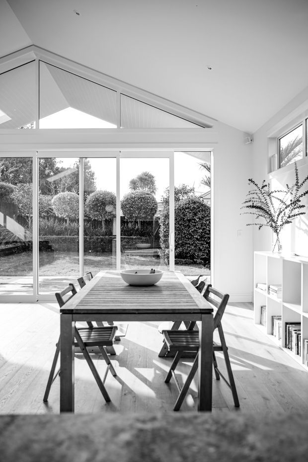 Bright monochrome photograph of a dining area with a solid timber dining table and folding chairs, positioned beside expansive floor-to-ceiling glazed sliding doors. The room has pale timber flooring, white walls and ceiling, a high angled glazed opening, and a low white built-in shelving unit. A landscaped garden is visible beyond the glazing, while strong sunlight creates defined shadows across the floor.
