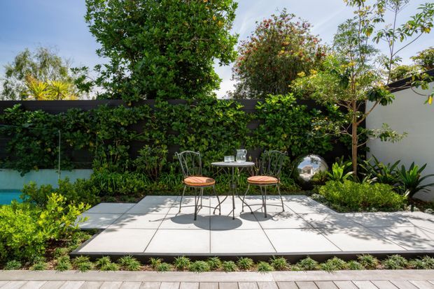 A landscaped courtyard garden with a small paved outdoor seating area, featuring a round metal café table and two chairs with coral seat cushions. Lush layered planting, a dark boundary wall with climbing greenery, a turquoise pool or water feature at left, and a reflective garden sphere create a private, resort-like setting.