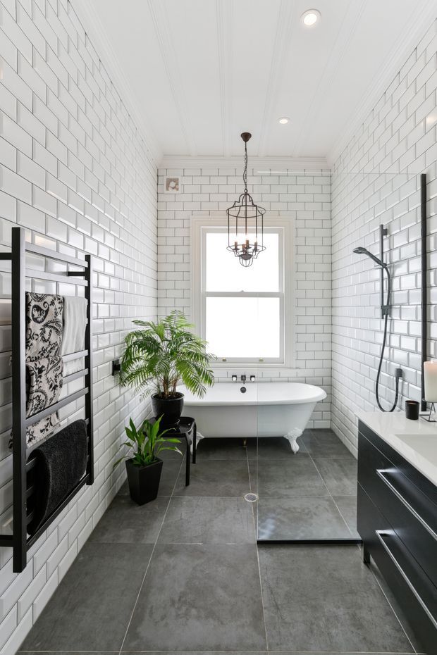 Bright, narrow contemporary bathroom with white subway-tiled walls, dark grey tiled flooring, a freestanding white clawfoot bathtub beneath a window, black-framed shower fittings, dark vanity cabinetry, a heated towel rail, potted plants, and a decorative pendant light. The room has a clean, monochromatic black-and-white palette with traditional detailing in the tub and ceiling trim.