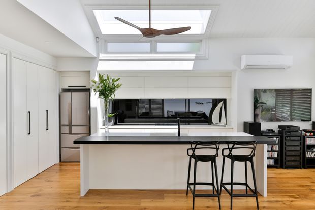 Bright open-plan kitchen and living area with a large white island, integrated cabinetry, stainless-steel refrigerator, black undermount sink and tap, and two black bar stools. White built-in joinery surrounds a dark reflective appliance or television niche, while timber flooring, a skylight, clerestory glazing, and a sculptural timber ceiling fan add warmth and natural light. A living area with media shelving is visible to the right.