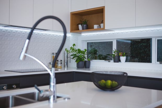 Contemporary kitchen with a large white island and integrated sink, dark lower cabinetry, white upper cabinetry, a textured geometric splashback, under-cabinet lighting, timber open shelving, and a wide window above the benchtop. Potted plants and a fruit bowl add natural accents.