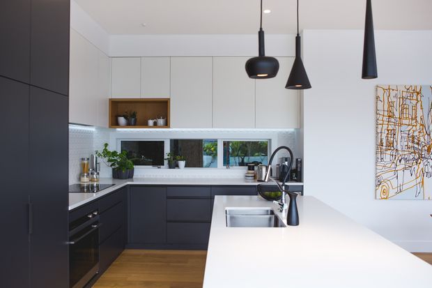 Contemporary kitchen with dark flat-panel cabinetry, white overhead cupboards, a large white island with integrated sink, timber flooring, patterned tile splashback, and black sculptural pendant lights. Indoor plants and artwork add restrained decorative detail.