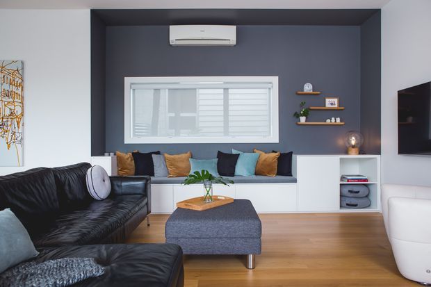 Contemporary living room with a dark charcoal feature wall, built-in window seat and storage, floating timber shelves, black leather sectional seating, and a large upholstered ottoman. White walls, warm timber flooring, colourful cushions, and recessed wall-mounted air conditioning create a clean but inviting interior.