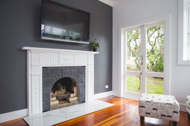Bright living room featuring a restored white decorative mantel and dark brick fireplace surround, a wall-mounted television, dark charcoal feature wall, polished timber flooring, and a glazed exterior door opening to a garden. A patterned ottoman sits in the foreground.