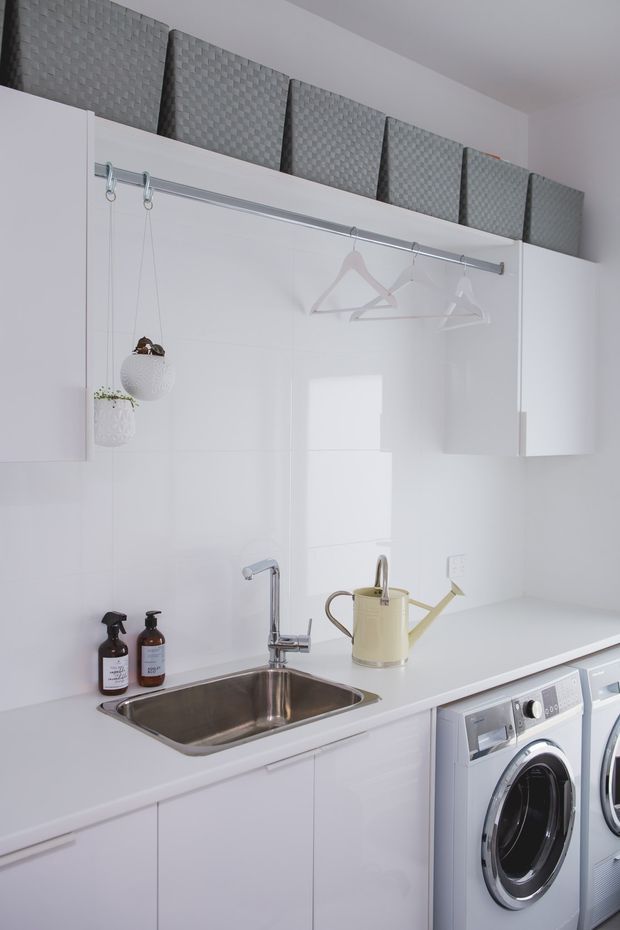 Bright, compact white laundry room with integrated cabinetry, a stainless-steel sink and chrome tap, front-loading washing machines, a long hanging rail with empty hangers, and grey woven storage baskets above the cabinets. Small hanging plants and a pale yellow watering can add subtle decorative accents.