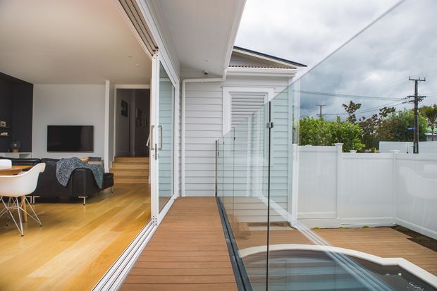 A contemporary residential indoor-outdoor living area opens through large sliding glass doors onto a timber-look deck beside a small plunge pool or spa. The exterior has white horizontal weatherboard cladding, a white privacy fence, and frameless glass balustrading. Inside, a warm timber floor, dark feature wall, television, sofa, and dining furniture are visible. Overcast coastal-looking conditions and neighbouring utility infrastructure appear in the background.