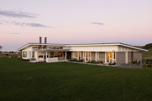 Single-storey contemporary home set in a broad landscaped lawn, photographed at dusk. The residence has a long low-pitched roof with deep eaves, extensive glazing, white vertical cladding and trim, horizontal grey wall cladding, a sheltered covered outdoor living area, and twin dark metal chimneys. Open rural fields are visible beyond the house.