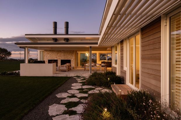 Twilight exterior view of a contemporary single-storey home with horizontal timber cladding, extensive timber-framed glazing, and a prominent slatted pergola or roof overhang. A landscaped garden path of irregular stone pavers leads toward a covered outdoor living area with a fireplace and seating. Warm interior lighting creates an inviting connection between the house and garden.