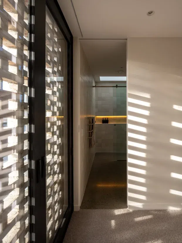 A narrow, modern interior passage leading to a tiled bathroom or shower area. A full-height black-framed glazed door or window runs along the left side beside a textured concrete breeze-block screen, casting strong horizontal bands of sunlight across the white walls and carpeted floor. At the rear is a walk-in shower with pale rectangular wall tiles, a skylight, dark flooring, a timber-toned shelf with concealed warm lighting, and a wall-mounted towel rail. The space has a minimalist, light-filled architectural character.