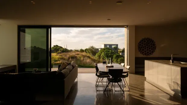 A warm, open-plan contemporary interior viewed from the living area toward a dining space and kitchen. Full-height black-framed glazing opens onto a natural coastal-looking landscape and floods the polished concrete floor with late-afternoon light. The room includes dark sofas, a compact dining table with black wire-frame chairs, white kitchen cabinetry with a dark benchtop, recessed ceiling lighting, and a circular geometric wall feature.