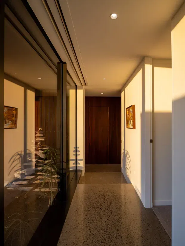 A narrow, warmly lit residential hallway or entry corridor with polished speckled flooring, white walls and ceiling, dark timber doors and feature joinery, and full-height black-framed glazing or mirrored panels along the left side. Recessed downlights, artwork, and reflected indoor planting add detail. Strong late-afternoon sunlight enters from the right, creating long shadows and a calm, refined atmosphere.