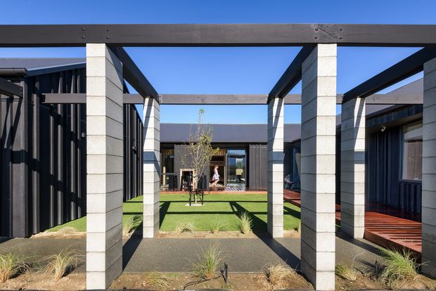 A contemporary single-storey architectural courtyard viewed along a paved external walkway. Dark vertical cladding and exposed black steel beams frame pale masonry columns, creating a strong pergola-like structural rhythm. The courtyard contains low ornamental grasses, a small tree, synthetic green lawn and timber decking, with surrounding glazed and solid building wings visible beyond.