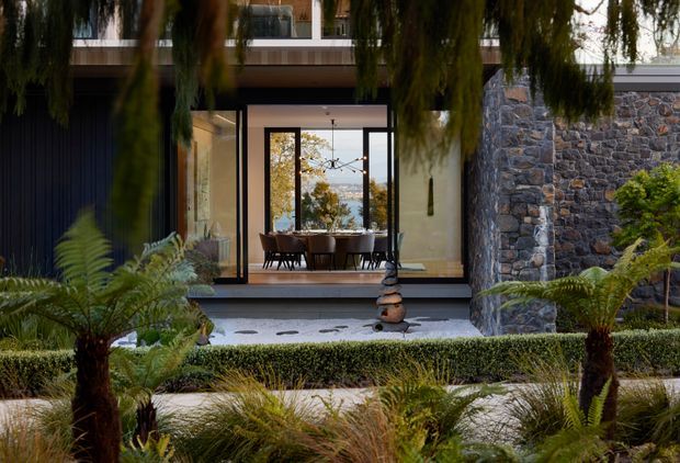 A landscaped courtyard approach frames a contemporary house with expansive dark-framed glazing, a dining area visible inside, and a prominent rough natural-stone wall. Ferns, clipped hedges, ornamental grasses, and a shallow stepping-stone water feature create a lush, tranquil entry garden.
