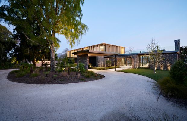 Wide exterior view of a contemporary single-storey residence set within a landscaped garden. The home features dark stone-clad walls, extensive glazing, a low horizontal roofline, and a prominent timber-clad upper roof volume with deep eaves. A covered entry or carport-like structure projects toward a pale curved driveway, with mature trees and ornamental planting surrounding the building.