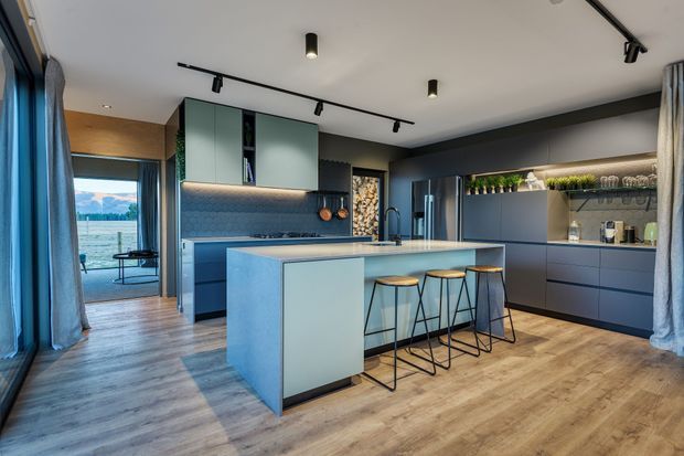 A spacious contemporary kitchen with a large central island and three stools, dark blue-grey cabinetry, pale green upper cabinets, integrated open shelving with plants, and a textured blue-grey splashback. Track lighting runs across the white ceiling, while warm timber-look flooring and full-height curtains frame glazed doors opening to an outdoor view.