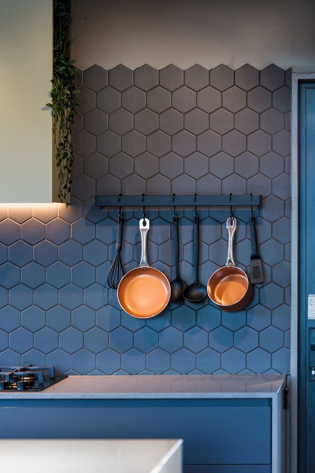 Close-up of a moody, contemporary kitchen featuring a full-height blue-grey hexagonal tile splashback, blue cabinetry, a pale benchtop, and a dark wall-mounted rail displaying copper pans and black utensils. A gas cooktop is partially visible at lower left, with a trailing indoor plant adding a natural accent.