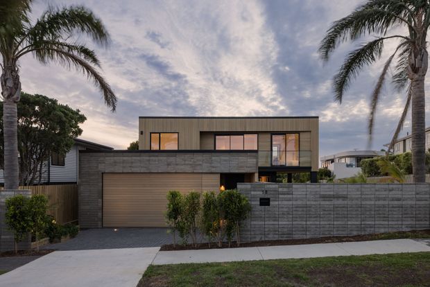 Street-facing view of a contemporary two-storey residence at dusk, with a rectilinear upper-level form, vertical timber-toned cladding, large rectangular windows, dark masonry walls, an integrated double garage, and a low concrete-block boundary wall. Tall palm trees and neighbouring homes frame the suburban setting.