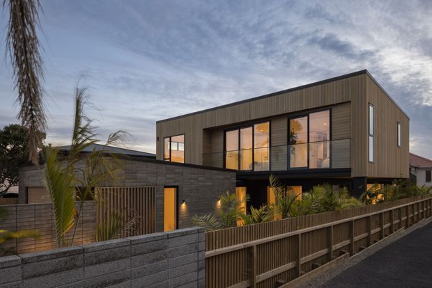 Exterior view of a contemporary two-storey residence at dusk, featuring a simple box-like form with a low-pitched or flat roof, dark vertical timber-look cladding, extensive black-framed glazing, and a lower level faced in dark masonry block. A timber slat fence, masonry boundary wall, landscaped palms, and warm interior lighting frame the street-facing facade.