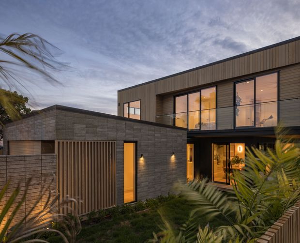 Exterior architectural photograph of a contemporary two-storey residence at dusk. The home combines dark horizontal masonry and vertical timber-look cladding, with extensive floor-to-ceiling glazing, recessed balconies, warm interior lighting, and a separate low flat-roofed garage or pavilion. Tropical planting and timber slat screening frame the foreground.