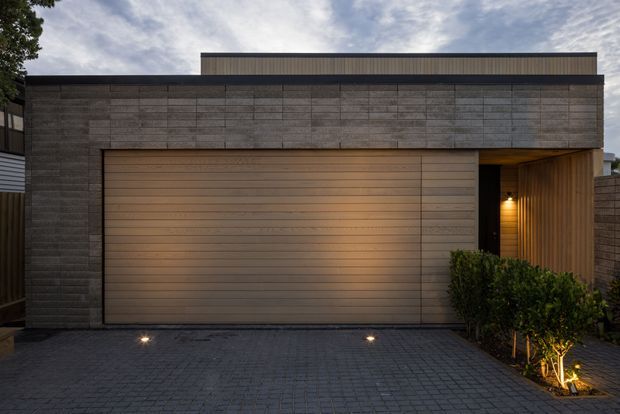 Street-facing view of a contemporary residential garage facade at dusk. The composition is dominated by a broad timber-toned sectional garage door set within a dark, horizontally articulated masonry or concrete-block facade. A recessed timber-lined entry alcove with warm lighting sits to the right, beside a neatly trimmed hedge. The driveway is paved in dark rectangular setts, with subtle integrated ground lighting and a flat, layered roofline.