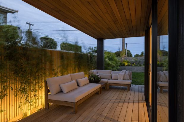 A covered outdoor lounge terrace with built-in timber decking, a warm timber-lined ceiling, dark structural posts, and a timber privacy screen. Cushioned lounge seating surrounds a low coffee table, with garden planting and a residential suburban setting beyond. Large dark-framed glazed doors connect the terrace to the interior.