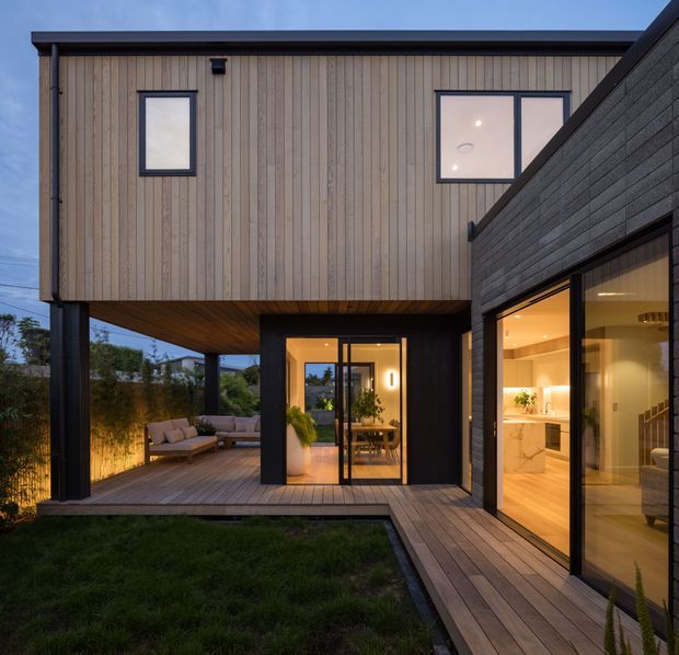 Twilight exterior view of a contemporary two-storey home arranged around a small landscaped courtyard. The upper level is clad in vertical light timber boards, while the lower volume features dark masonry cladding and large black-framed glazed openings. A timber deck and covered outdoor seating area connect the courtyard to the warmly illuminated kitchen and dining spaces inside.