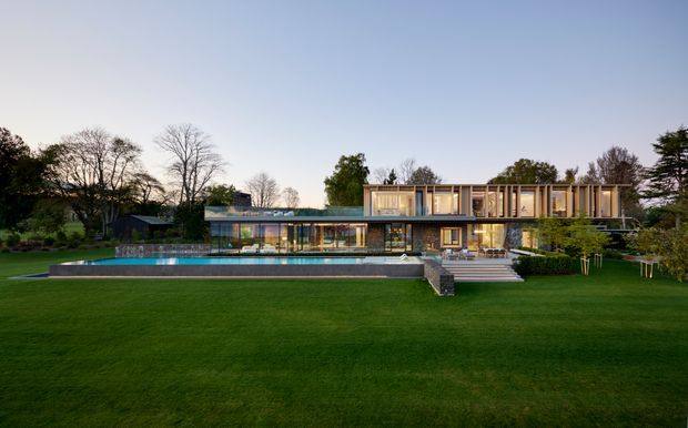 Wide exterior view of a large contemporary multi-level residence set within a landscaped lawn and mature garden. The home features extensive floor-to-ceiling glazing, horizontal lines, a projecting upper level with vertical timber-like screens, and a long rectangular swimming pool in the foreground.