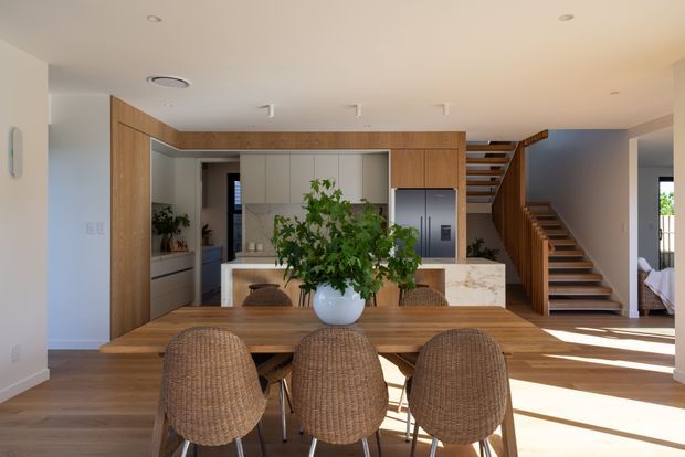 Bright open-plan dining and kitchen interior with a large solid-wood dining table and woven dining chairs in the foreground. The kitchen features light neutral cabinetry, a pale stone island, integrated stainless-steel refrigerator, and warm timber architectural framing. A timber staircase with open risers leads upward at the right, while indoor plants add a natural element. The room has a clean contemporary feel with abundant daylight and warm timber flooring.