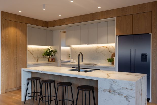 A warm contemporary kitchen with a large waterfall-edge island, four black bar stools, integrated cabinetry, a dark double-door refrigerator, undermount sink and black gooseneck tap. The kitchen features extensive light stone surfaces with subtle veining, pale grey upper cabinetry, and rich timber wall-height cabinetry and trim. Under-cabinet lighting creates a softly illuminated backsplash, while recessed ceiling lights provide balanced ambient lighting.