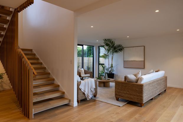 Warm contemporary open-plan living area with a timber staircase and vertical timber balustrade at left, opening onto a softly furnished lounge. The room features pale timber flooring, white walls and ceiling, woven rattan seating, a neutral sofa, indoor palms, recessed downlights, large black-framed glazed doors or windows, and restrained abstract wall art.