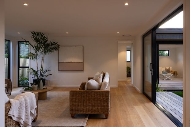 A warm, contemporary living room with pale timber flooring, neutral walls, recessed ceiling lights, and a large woven rattan sofa facing away from the camera. A soft area rug, white textiles, indoor palms, and understated wall art create a relaxed natural feel. Full-height black-framed glazing opens onto a timber deck and a small planted courtyard, while a hallway leads to additional rooms beyond.