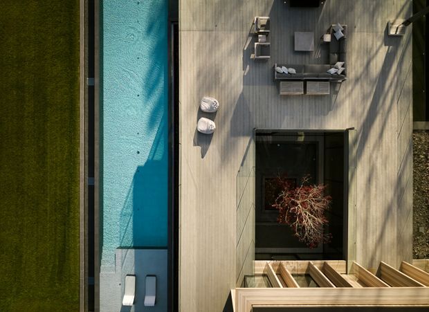 Aerial view of a contemporary outdoor terrace with a long narrow swimming pool beside a pale timber deck. The deck contains outdoor lounge seating, a central fire feature or sunken courtyard opening, and a stair or louvred timber structure at the lower edge. Strong linear geometry and tree shadows create a calm, modern composition.