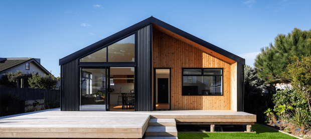 Exterior view of a compact contemporary gable-roof house with a raised timber deck and central entry steps. The facade combines dark vertical metal cladding with warm vertical timber boards, extensive black-framed glazing, and a glazed gable end. Landscaping surrounds the building, with a visible open-plan interior through the glazing.