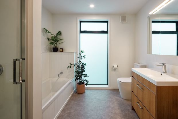 Bright contemporary bathroom viewed from the shower area, featuring a freestanding-style bathtub with white vertical wall panelling, a timber vanity with white benchtop and integrated basin, toilet, frosted window, large mirror, recessed ceiling light and linear vanity lighting. A potted plant adds a natural accent.