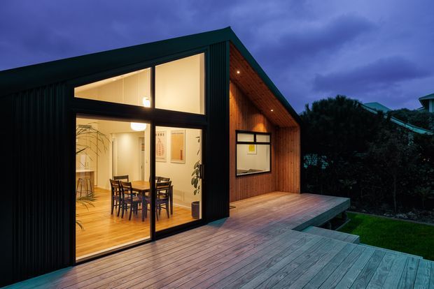Twilight exterior view of a compact contemporary pavilion or extension with a steep gabled roof, dark ribbed cladding, warm timber-lined gable, and extensive floor-to-ceiling glazing. The building opens onto a broad timber deck with steps leading to a landscaped garden. A warmly lit dining area is visible inside.