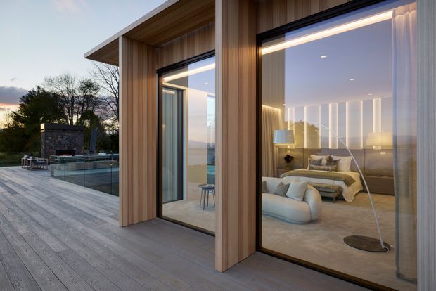 Contemporary single-storey pavilion viewed from an expansive timber deck at dusk. The facade features vertical timber cladding and large dark-framed floor-to-ceiling glazing, offering views into a warmly lit bedroom and adjoining interior. A glass pool barrier and outdoor fireplace area are visible to the left, with landscaped trees beyond. Inside, the bedroom has a bed, curved lounge chair, bedside lamps, pale carpet and softly illuminated vertical wall detailing.