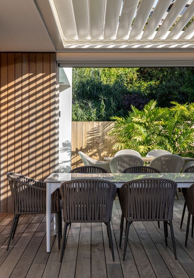 A covered outdoor dining area with a large rectangular table and woven dark chairs on a timber deck. The space is framed by vertical timber wall cladding and a white louvred roof or pergola, with lush tropical planting visible beyond a timber fence. Strong sunlight creates striped shadows across the wall, furniture, and deck.