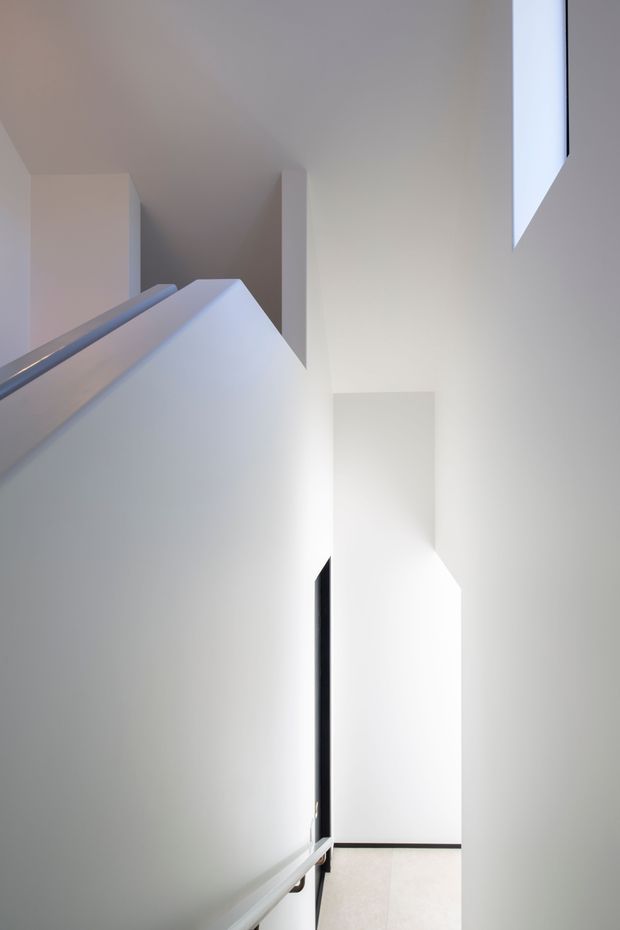 A minimalist interior stairway viewed upward, featuring crisp white walls, angular geometric forms, a white stair balustrade, and strong natural light creating soft shadows. A narrow glimpse of the lower landing and pale flooring is visible at the bottom.