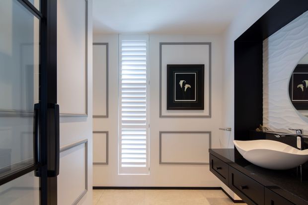 A refined contemporary bathroom featuring a freestanding sculptural basin on a dark built-in vanity, a black-framed mirror, and a textured white feature wall. The room has pale tiled flooring, white walls with decorative grey panel mouldings, a narrow plantation-shutter window, and black-framed glazing or cabinetry at the left.