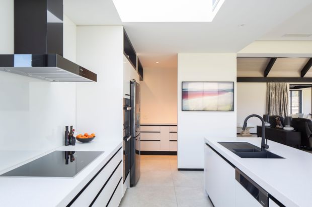 Bright contemporary kitchen with white flat-panel cabinetry, a large white island with integrated black sink and tap, black induction cooktop, dark integrated appliances, and a black-and-white rangehood. A skylight or large ceiling opening provides natural light. The kitchen opens toward a dining or living area with dark exposed ceiling beams and curtains.
