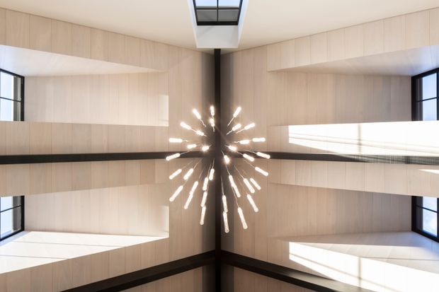 Upward-looking view of a tall, light-filled contemporary interior atrium or stairwell, with pale timber wall panelling, deep projecting horizontal openings, black-framed windows, and a dramatic radial pendant light suspended at the centre. A skylight brings daylight into the high ceiling.
