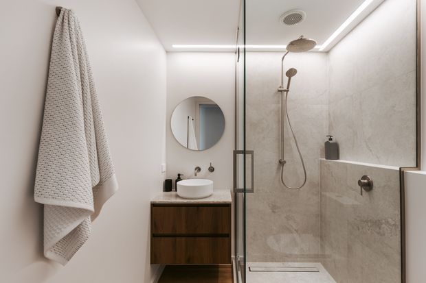 Contemporary compact bathroom viewed from the entry, featuring a frameless glass shower enclosure with rainfall showerhead and handheld fitting, large light stone-look wall tiles, a floating dark timber vanity with white vessel basin, round mirror, recessed ceiling lighting, and a textured white towel. The space has a clean, warm, refined appearance.