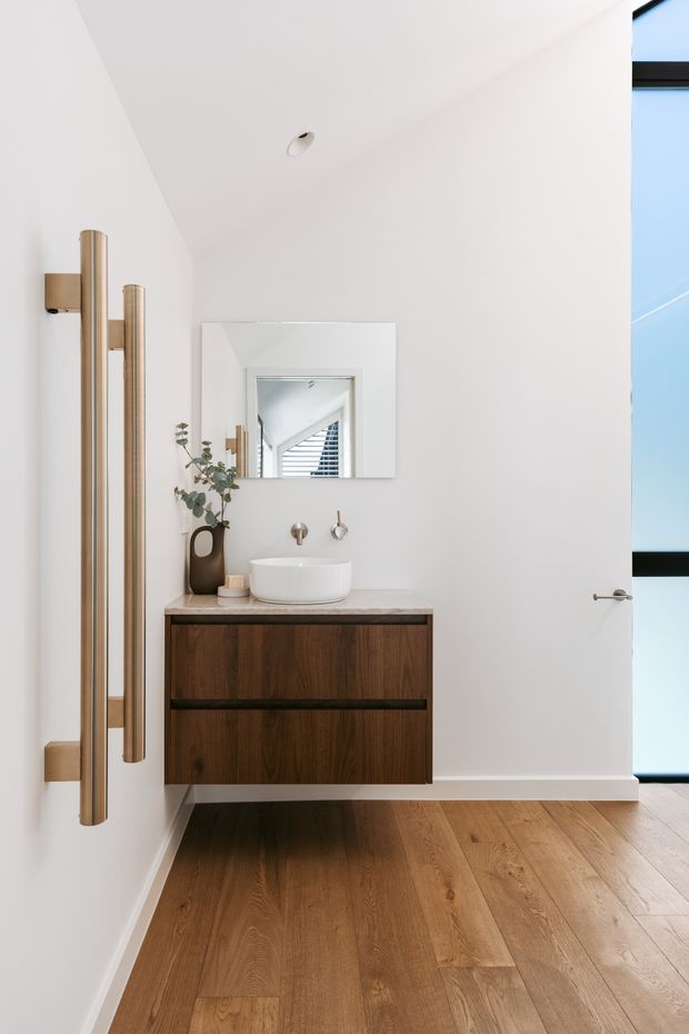 Bright, minimalist bathroom with a floating dark timber vanity, white vessel basin, wall-mounted brushed metal tapware, round mirror, and warm timber flooring. A large frameless glass door or partition is visible on the right, with a tall brushed metal heated towel rail on the left wall. The mirror reflects a stairway and louvred window beyond.