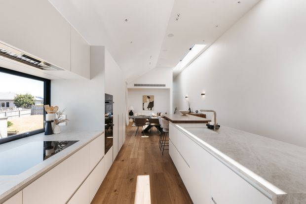 Bright, elongated contemporary kitchen with white handleless cabinetry, pale stone benchtops, integrated appliances, timber flooring, a central island, and a dining area beyond. Large side glazing and a roof skylight bring in abundant natural light, creating a clean, airy open-plan interior.