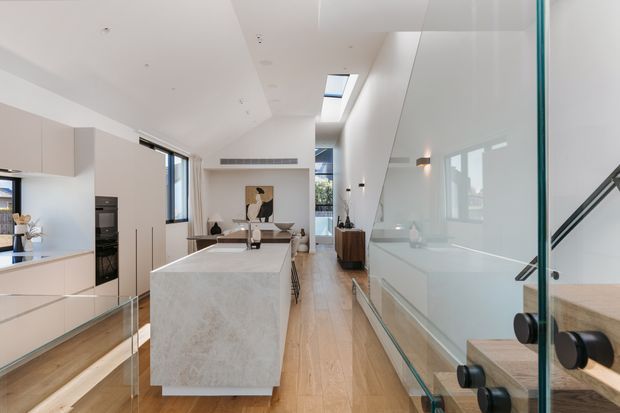 Bright contemporary open-plan kitchen and circulation area with a large pale stone island, light timber flooring, integrated pale cabinetry, skylights, and a glass-sided staircase with black metal detailing. A dining or living area is visible beyond the island.